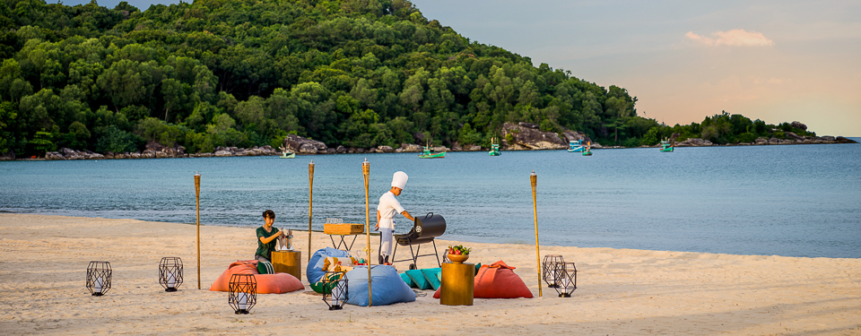 a man in a white chef hat tends to a barbecue next to the ocean while a woman decorates a small sitting table at the New World Phu Quoc Resort 