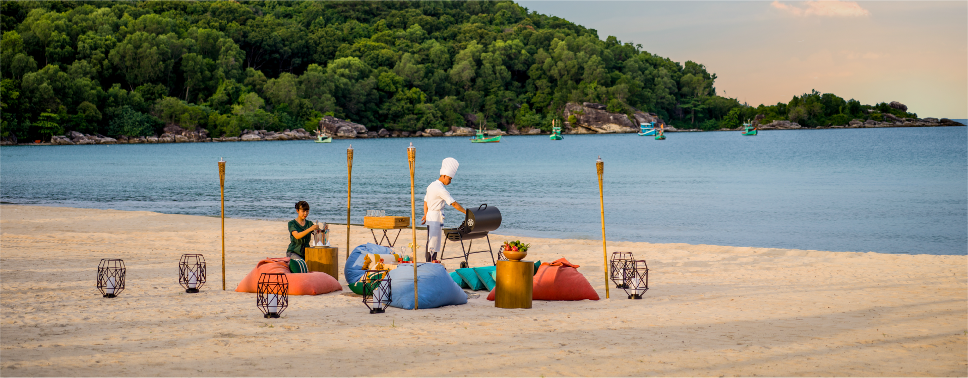 a man in a white chef hat tends to a barbecue next to the ocean while a woman decorates a small sitting table at the New World Phu Quoc Resort in Vietnam